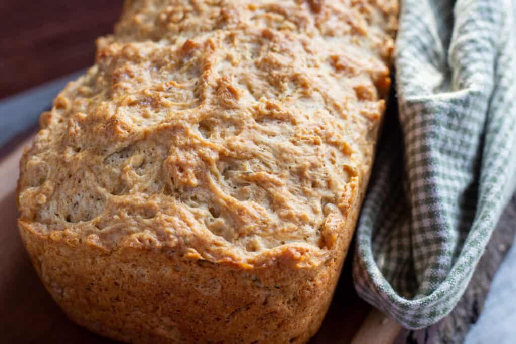 herbed beer bread sitting on a wooden board. To the right of the bread there is a green and beige checked cloth.