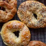 A variety of homemade bagels topped with sesame seeds, poppy seeds, everything bagel seasoning, shredded cheese sitting on a wire rack.