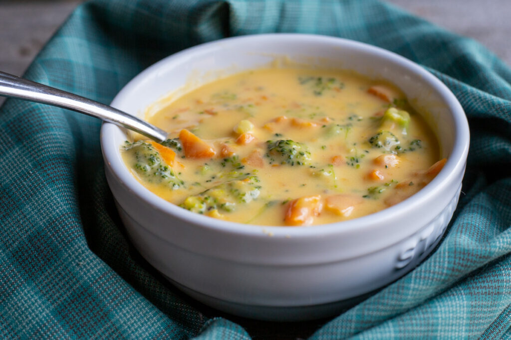 Broccoli Cheddar Soup in a white bowl with a spoon to the left of the bowl. The bowl is surrounded by teal coloured napkin.