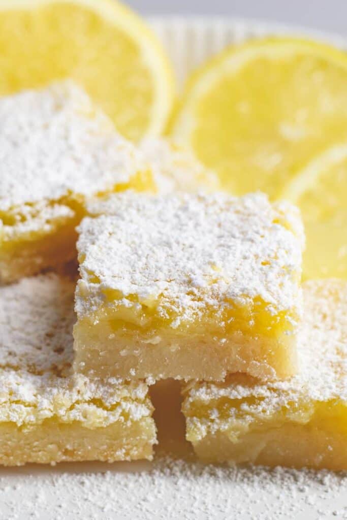 Lemon bars stacked on marble board with sliced lemons in the background.