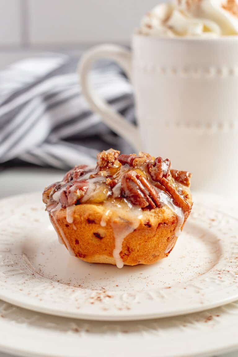 Cinnamon roll apple pie cup on white plate with coffee in the background.