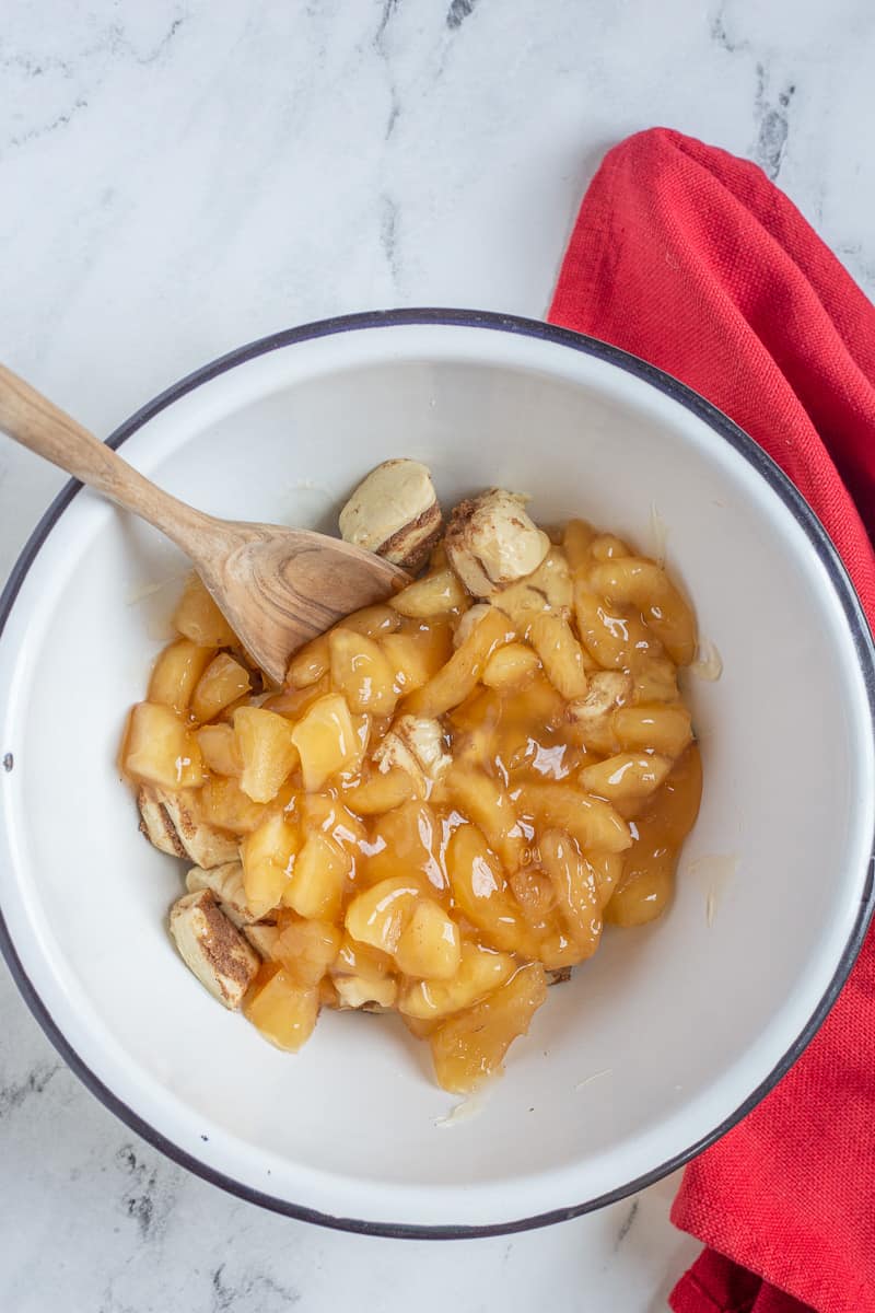 Apple pie filling and cut up cinnamon rolls in mixing bowl with spoon.