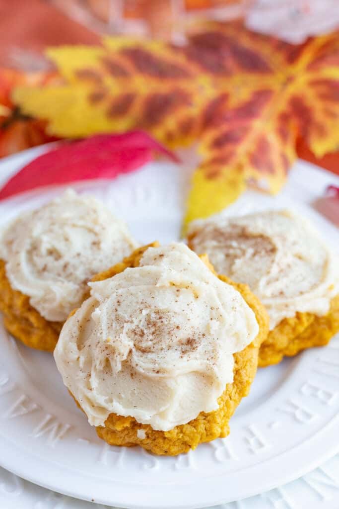 Pumpkin cookies with brown sugar frsoting on a white plate with maple leaves in the background.