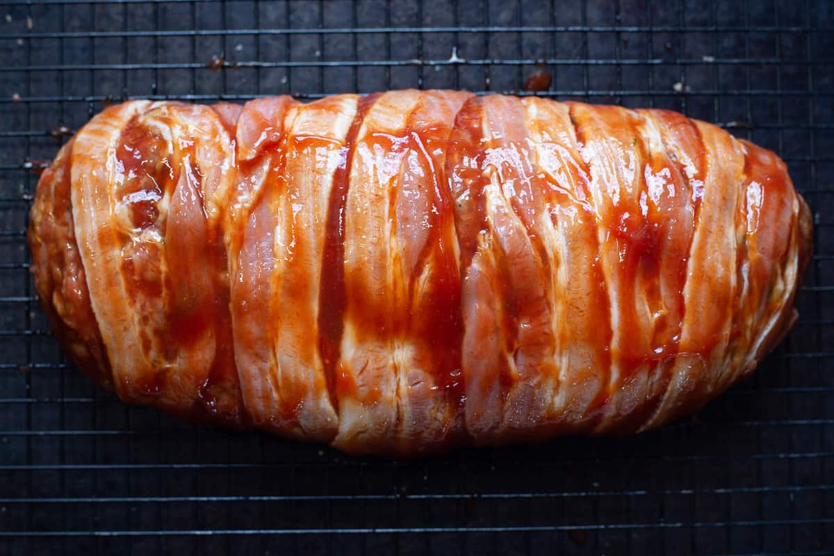 Bacon is wrapped around shaped meatloaf on wire baking rack ready to go into the oven.