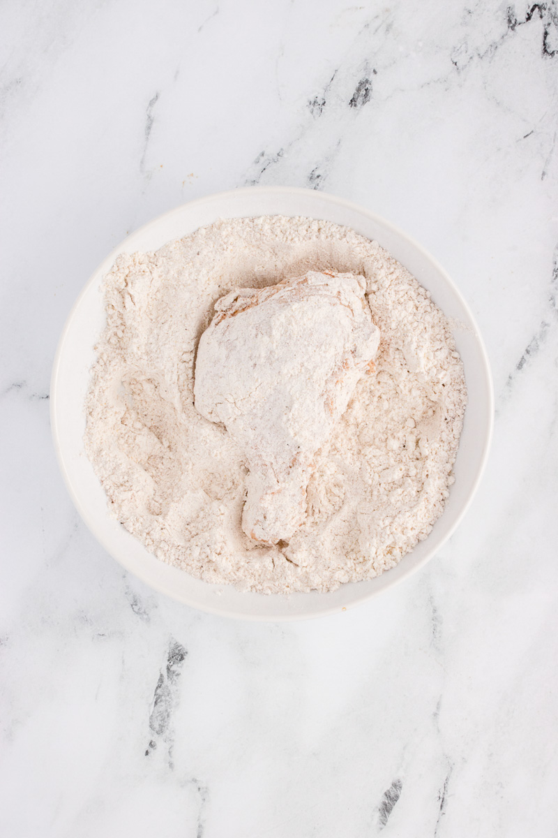 Chicken piece is dredged in flour in white bowl.