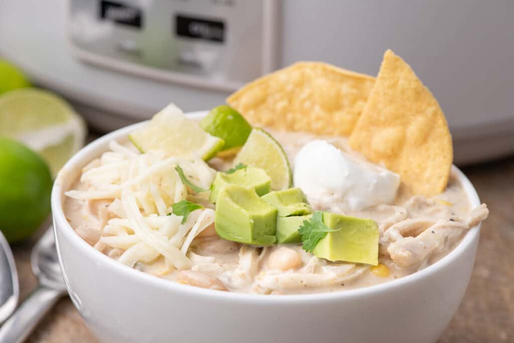 A bowl of creamy white chicken chili topped with shredded cheese, diced avocado, tortilla chips, a dollop of sour cream, and cilantro. A lime wedge is on the rim. A slow cooker is in the background.