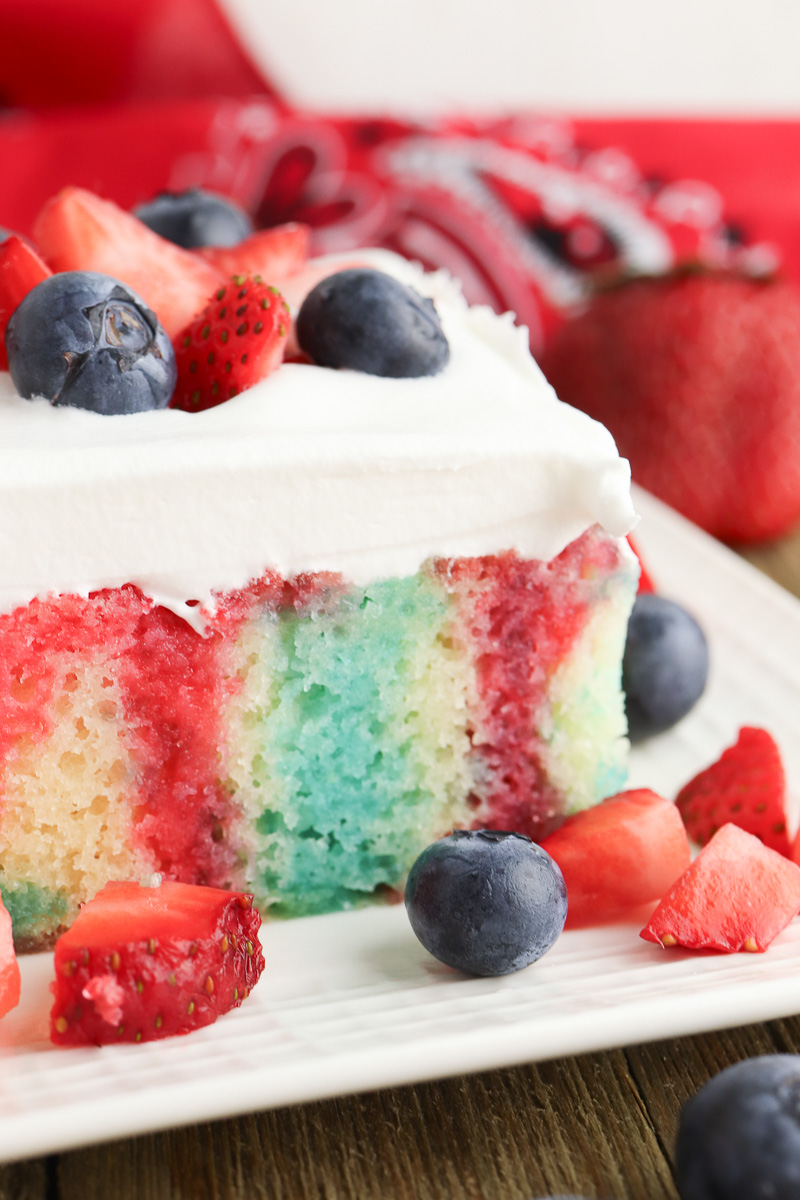 Piece of berry poke cake on a white plate with blueberries and strawberries around it.