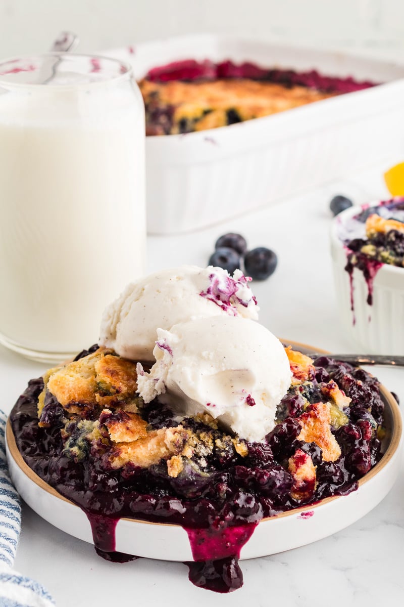 Blueberry dump cake with frozen berries and ice cream on top on a white plate.