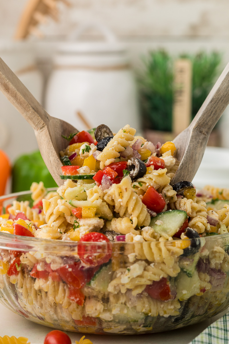 Greek pasta salad is scooped from a glass bowl with wooden spoons.