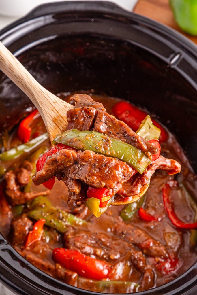 Crockpot pepper steak scooped up on a wooden spoon.