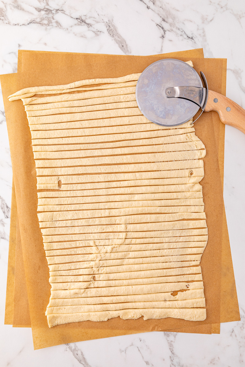 Crescent roll unrolled and cut into strips on cutting board with pizza cutter on the side.