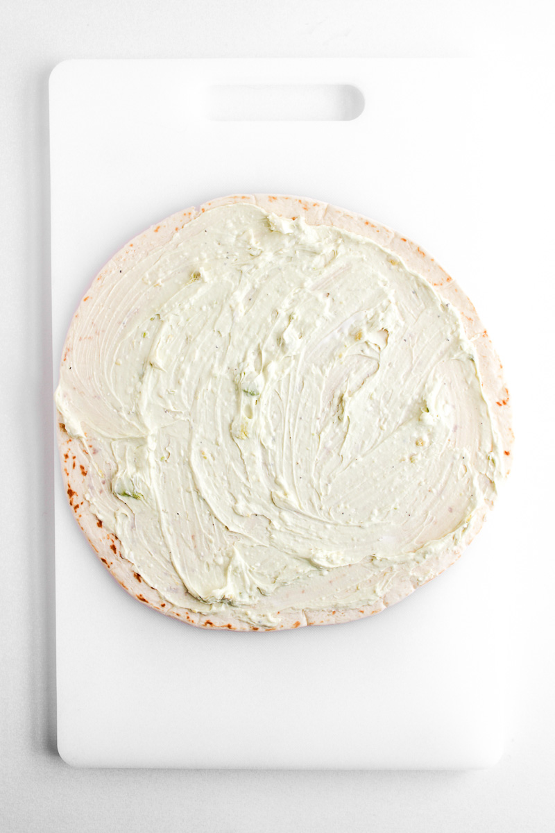 Avocado cream cheese mixture is sptread on flour tortilla.