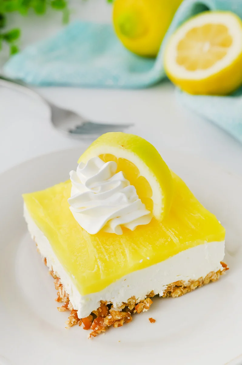 Piece of lemon pretzel salad on a white plate with whip cream and slice f lemon on top. 