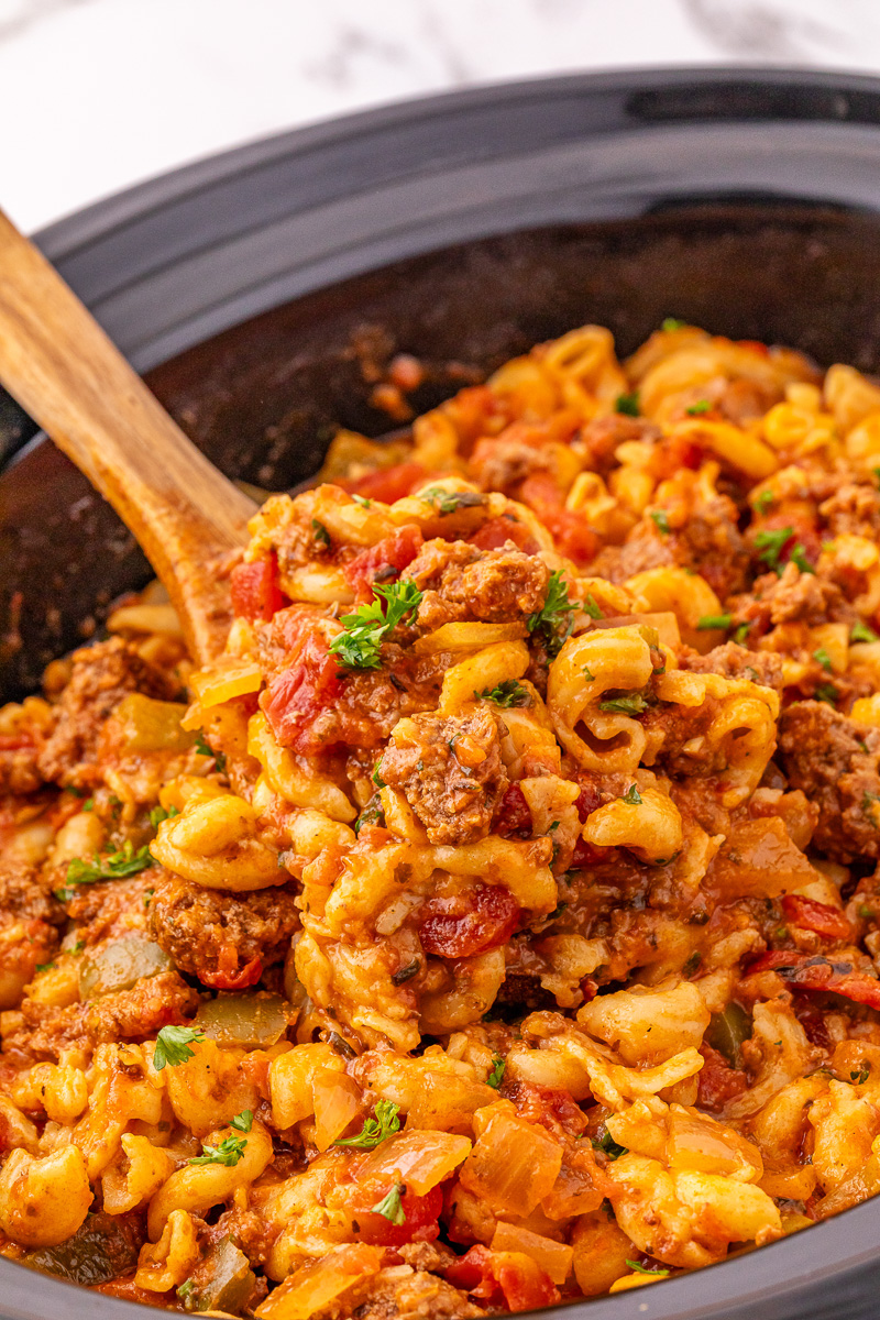 Scoop of American goulash held up over crockpot with remaining dinner.