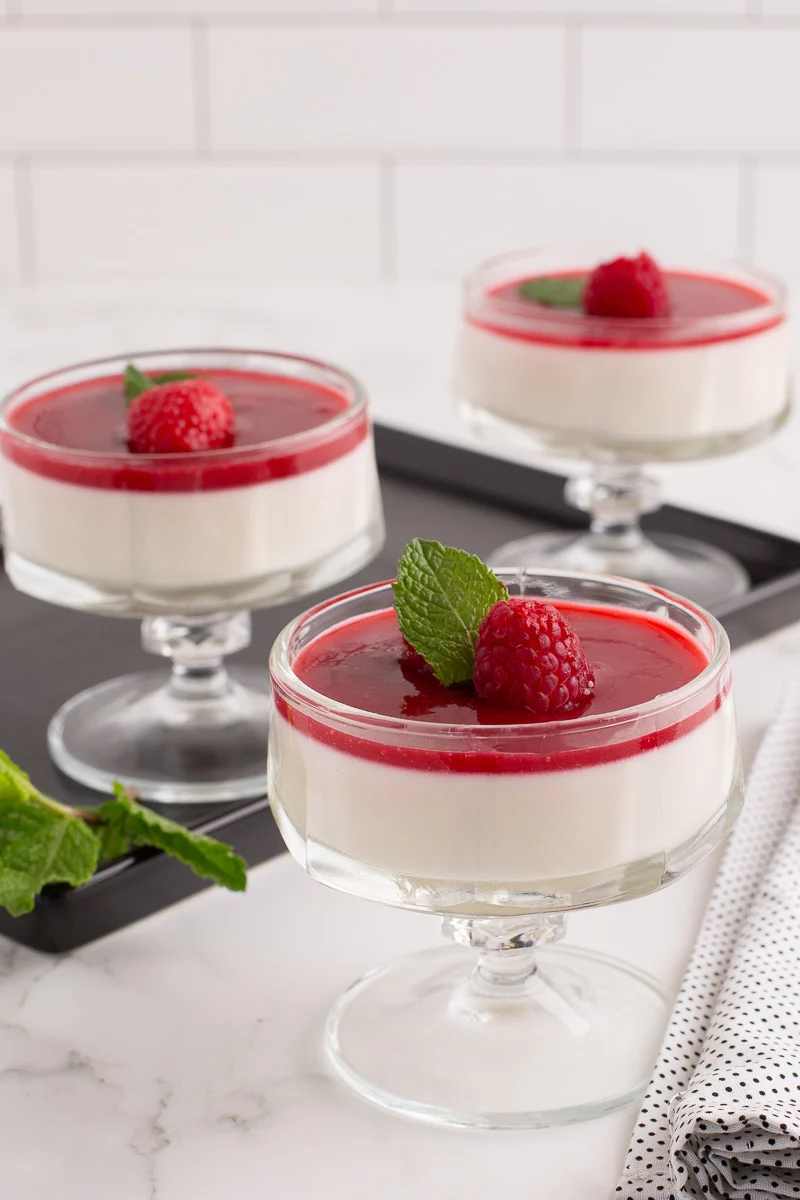 Panna cotta in glass serving bowls with a fresh raspberry and mint on top.