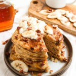 Banana bread pancakes stacked on a plate and topped with whipped cream and chopped pecans.