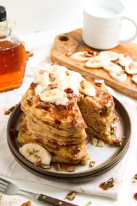 Banana bread pancakes stacked on a plate and topped with whipped cream and chopped pecans.