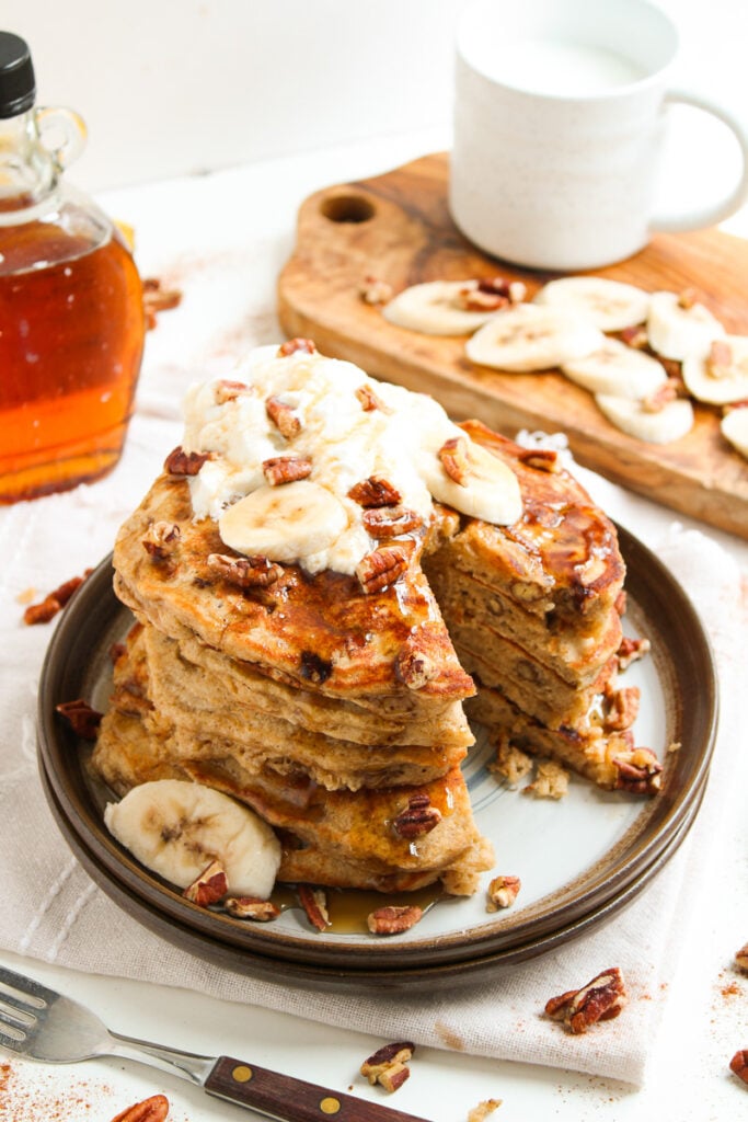 Banana bread pancakes stacked on a plate and topped with whipped cream and chopped pecans.