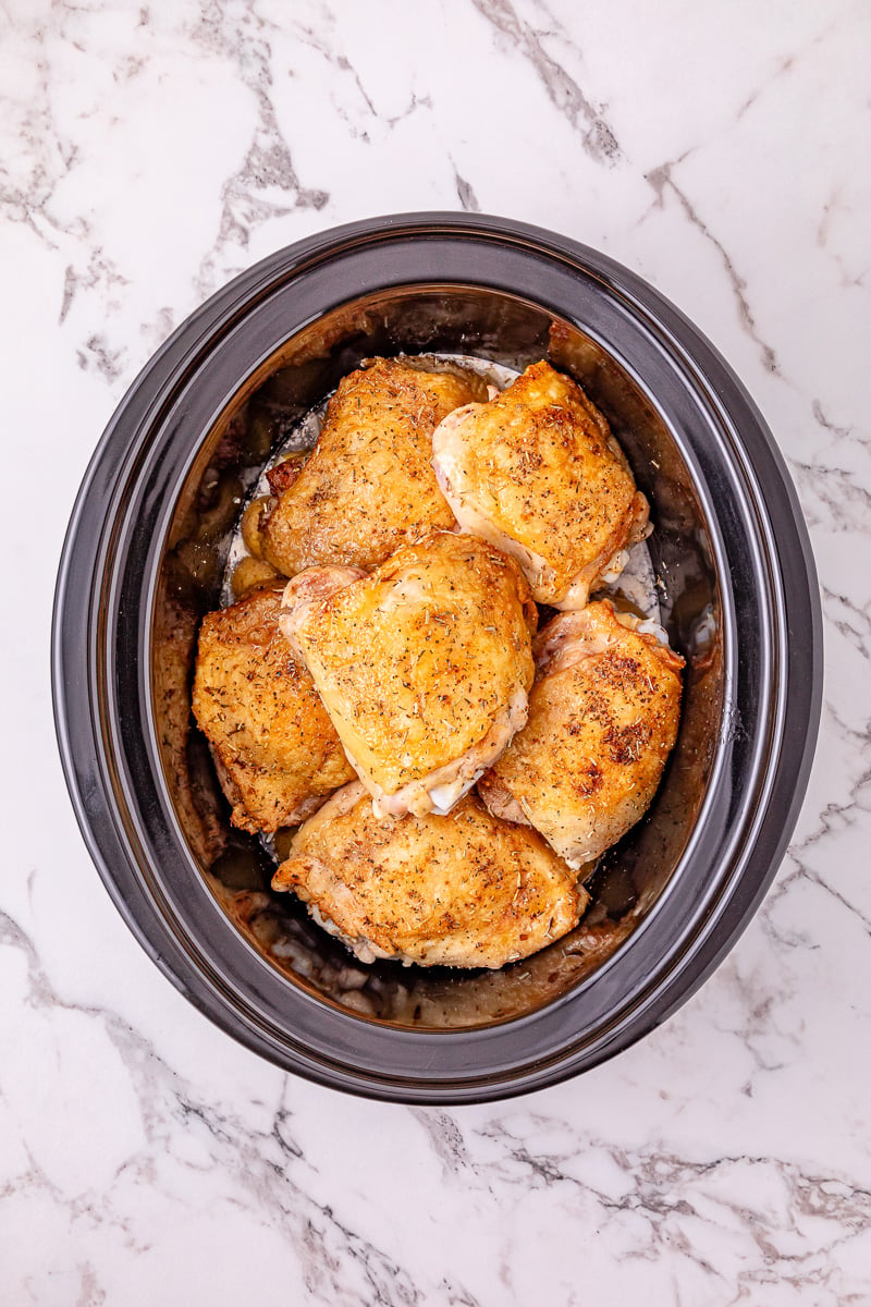 Seared chicken thighs are placed on top of potato mixture in crockpot.