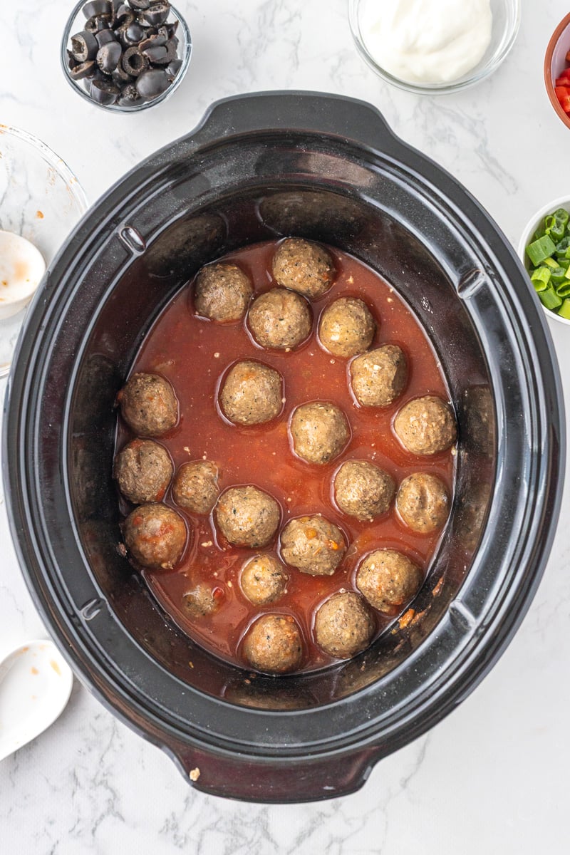 Enchilada sauce is poured over top of meatballs in slow cooker.