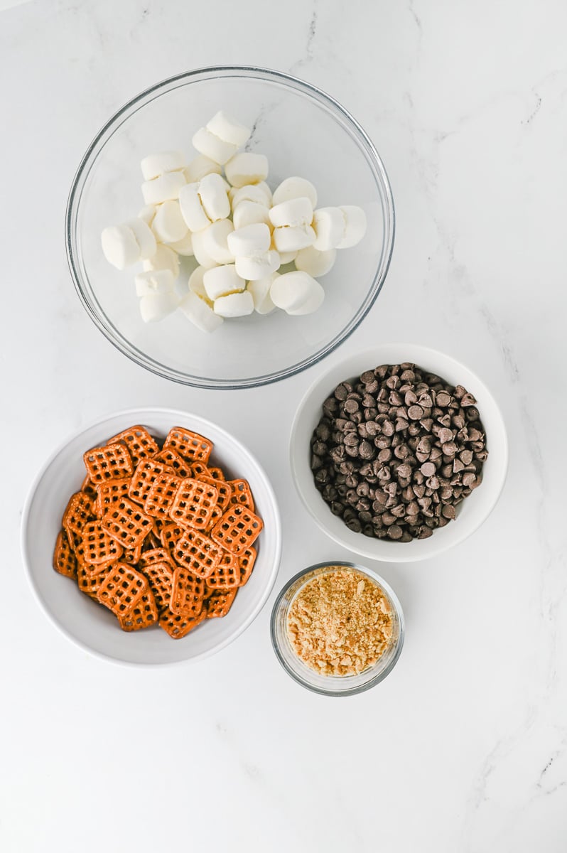 S'mores pretzel bites ingredients laid out in small bowls on kitchen counter.