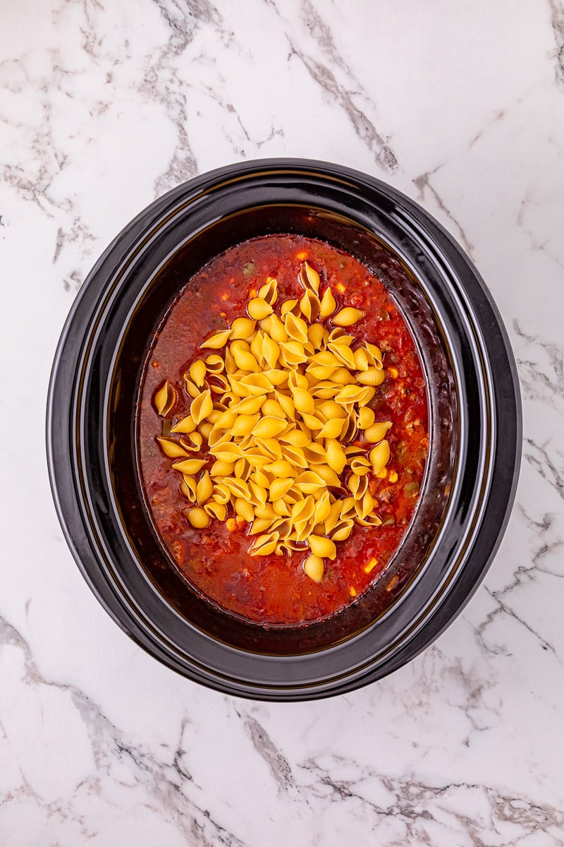 Uncooked pasta shells are stirred into meat mixture in crockpot.