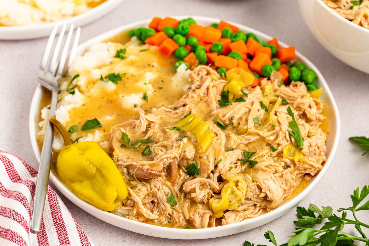 Mississippi chicken served on a plate with mashed potatoes, carrots, peas and au jus gravy.
