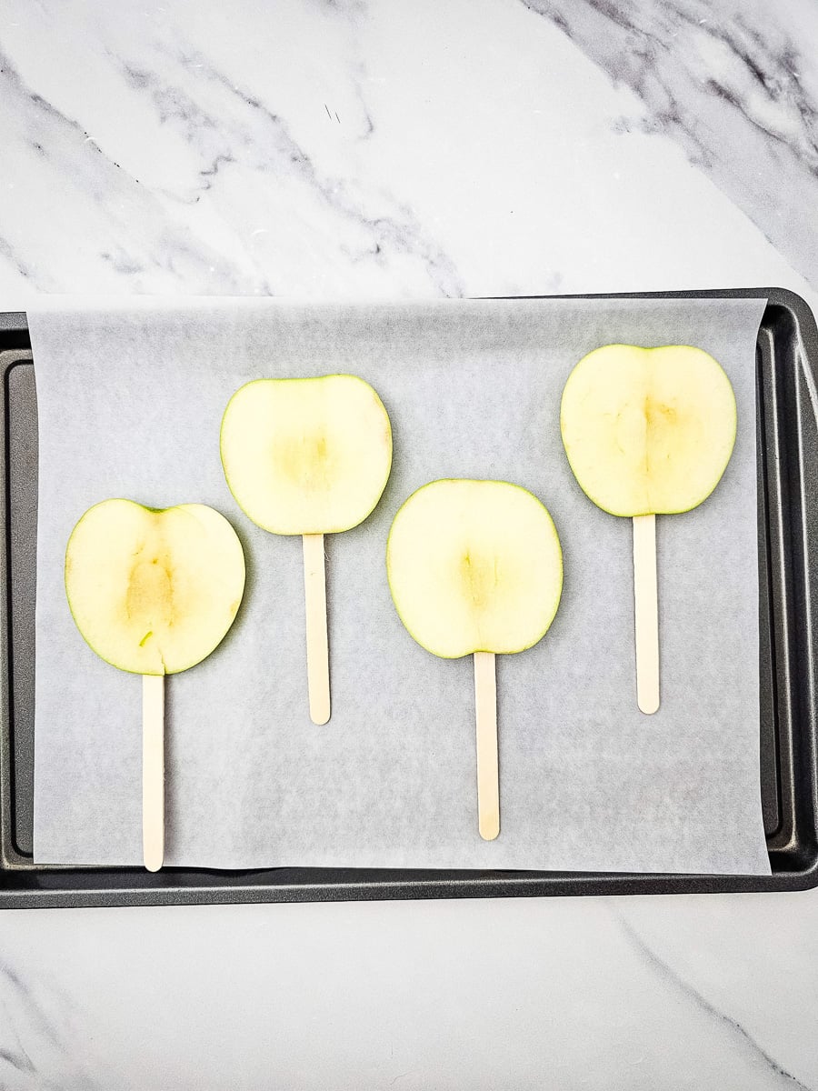 Sliced apple with cake pop stick in them on parchment lined baking sheet.