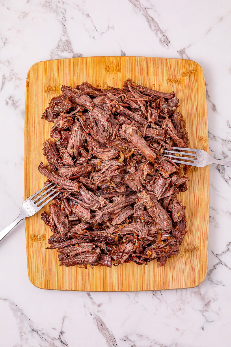 Beef is shredded with two forks on wooden board.