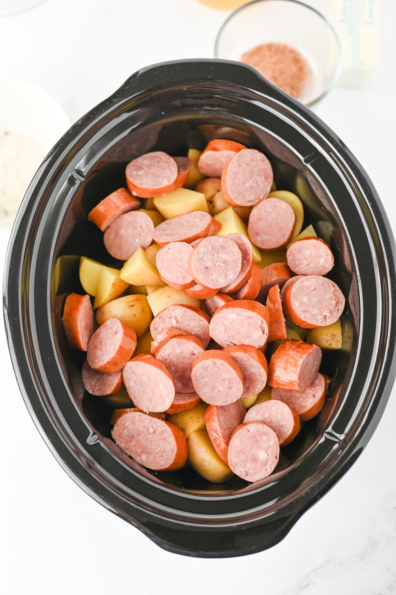 Sliced sausages are placed on top of potatoes in the crockpot.
