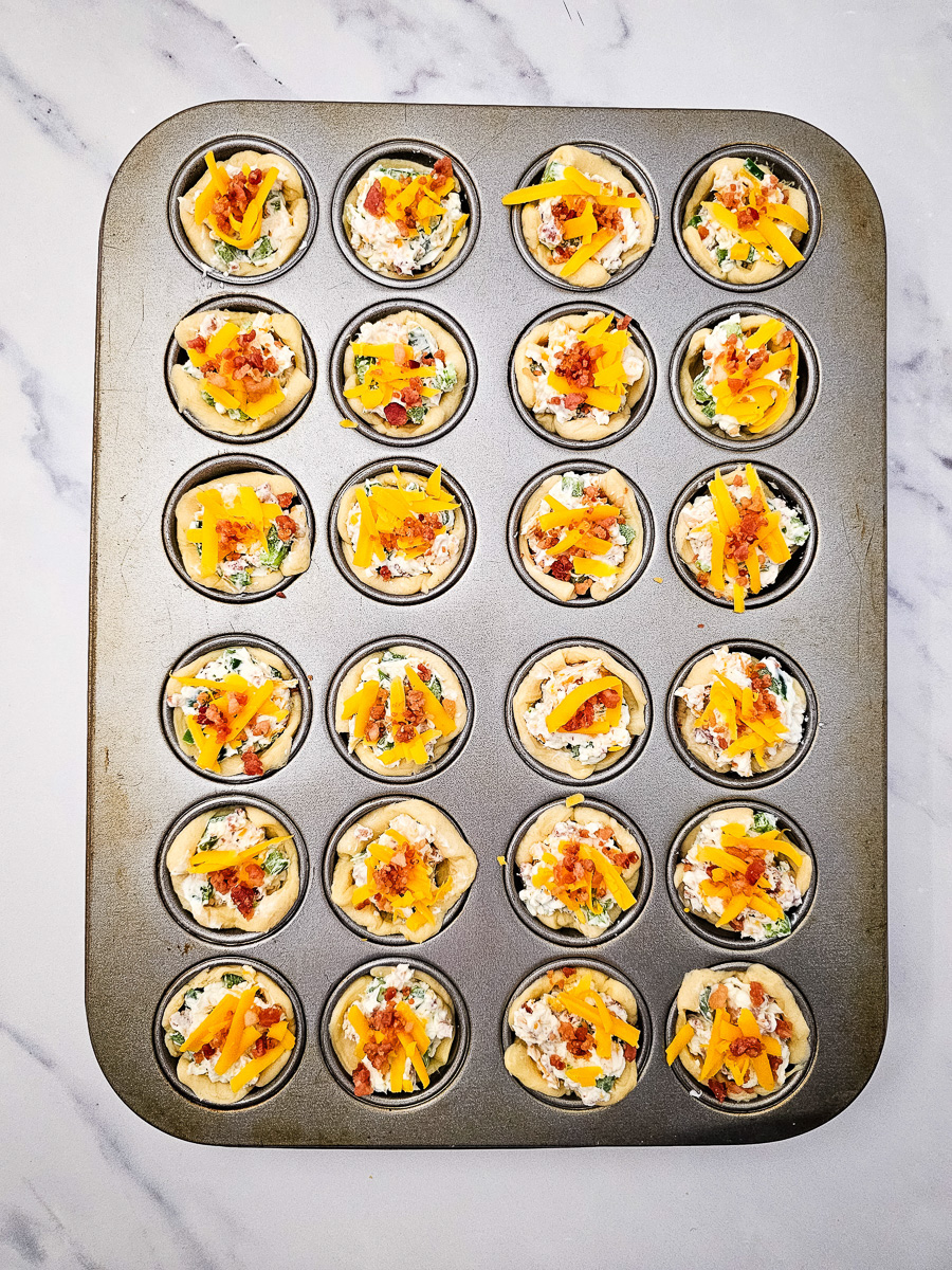 Cream cheese mixture, shredded cheese and bacon bites are sprinkled on top of each muffin tin.