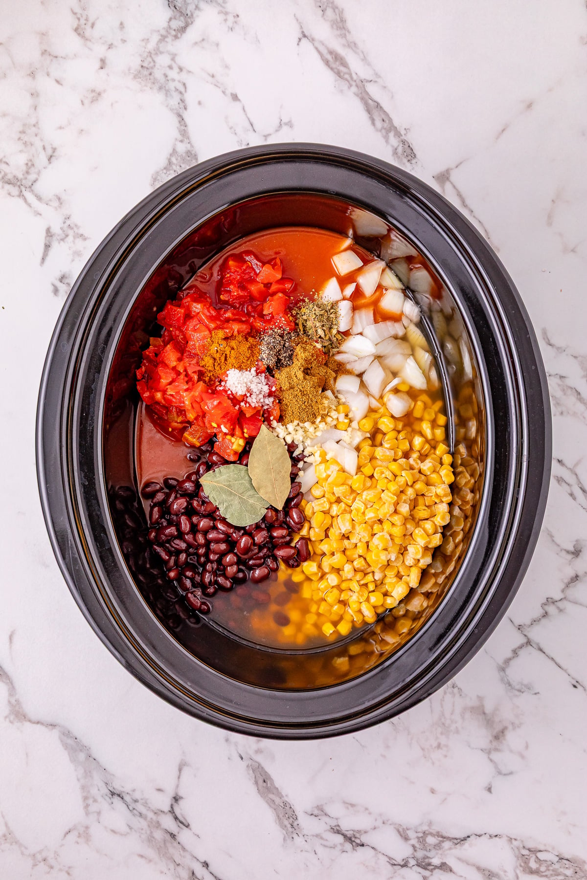 Onion, garlic, black beans, red enchilada sauce, diced tomatoes and green chilies, corn kernels, chicken broth, seasonings, and bay leaves are added to the slow cooker on top of the chicken.