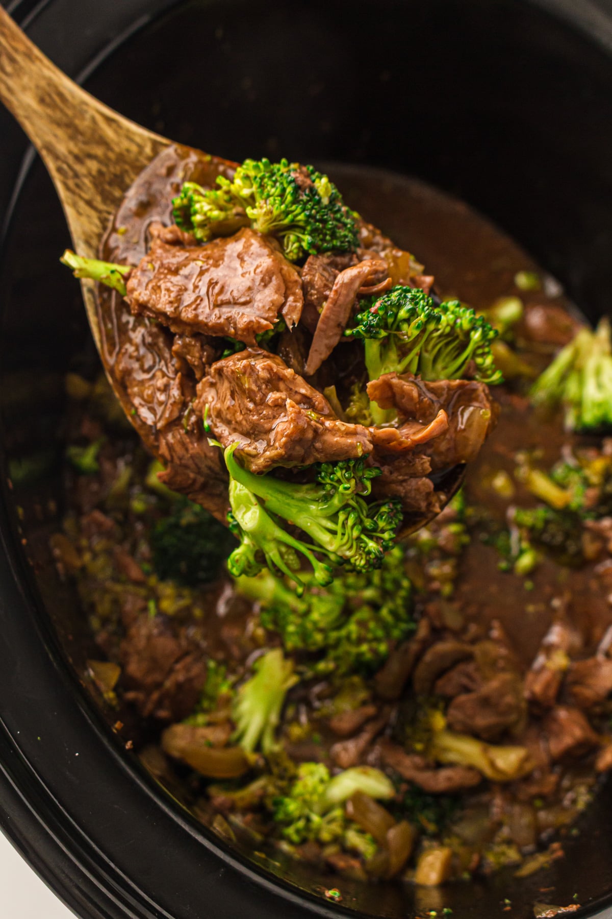 Beef and broccoli scooped up on a wooden spoon held over slow cooker.